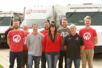 Smiling group of Weisser Distributing employees in Weisser Distributing apparel, standing in front of Weisser trucks.
